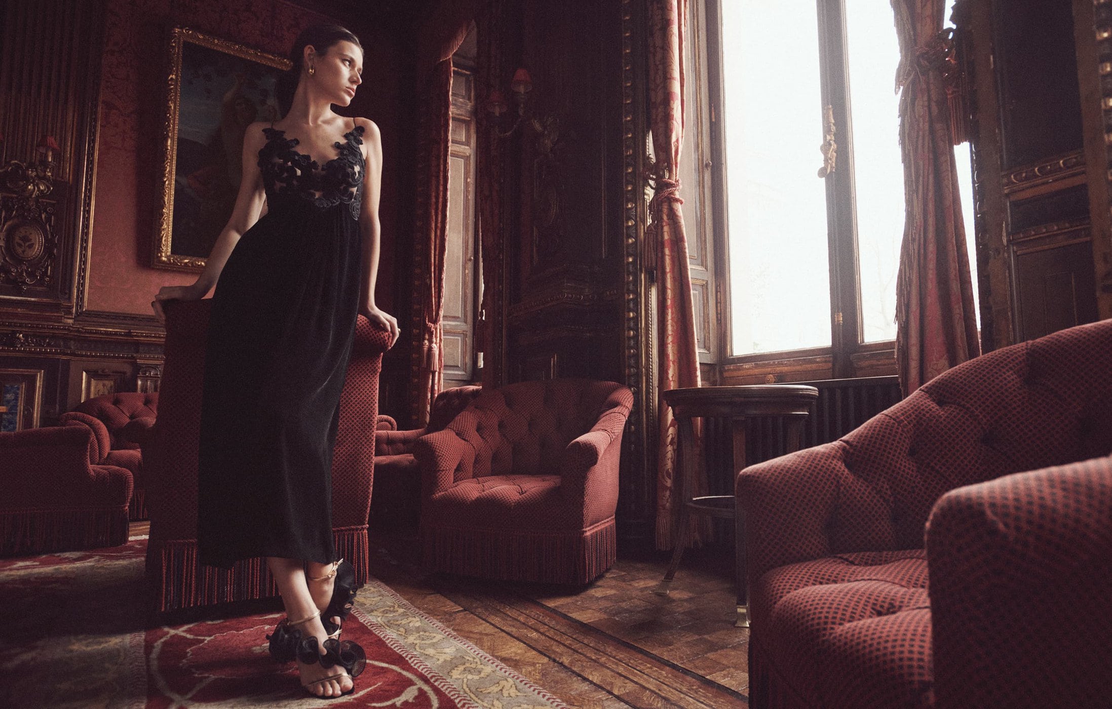 Elegant woman in a black dress posing in a vintage interior with red velvet armchairs, ornate walls, and soft window light.