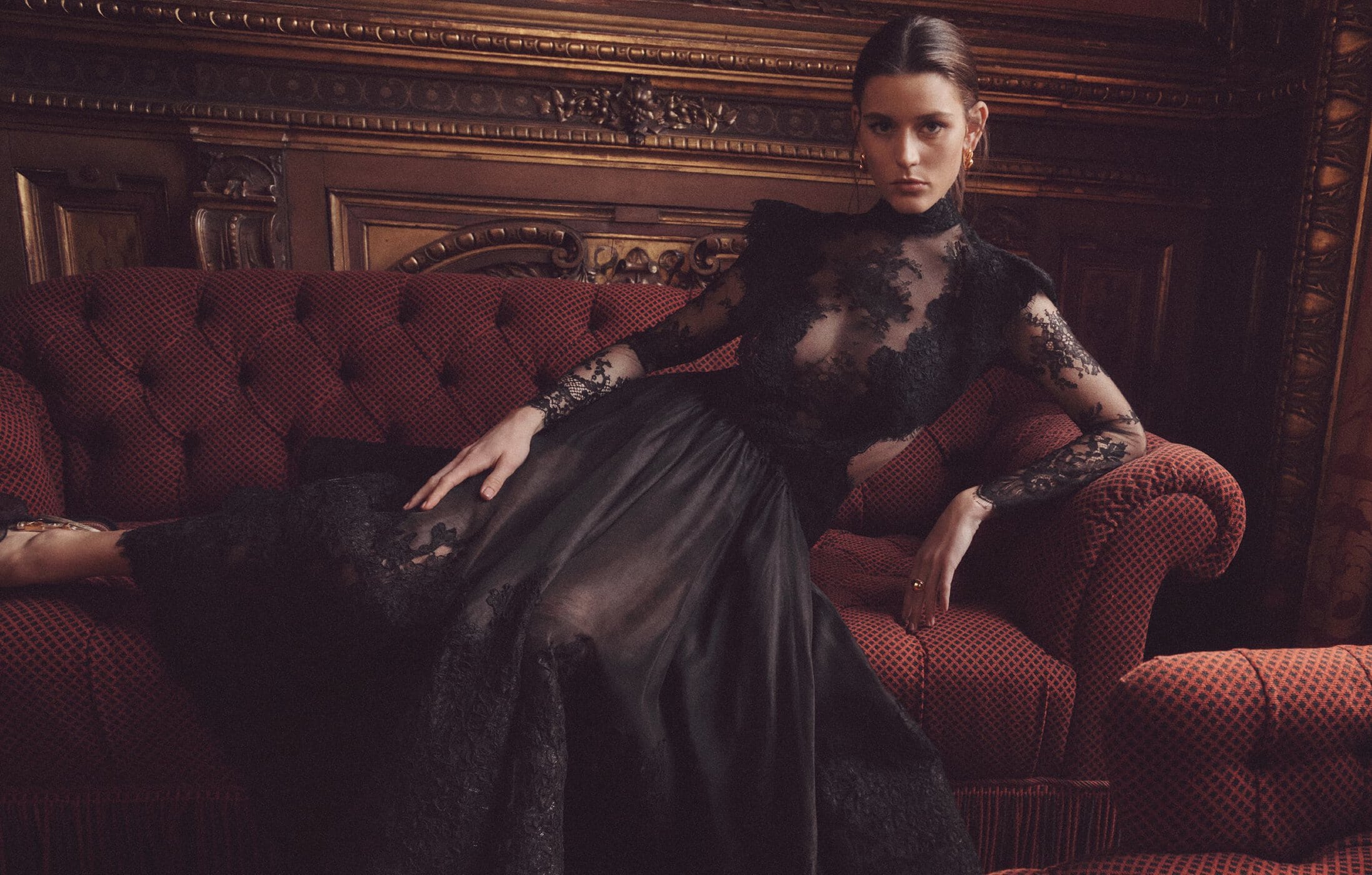 Brunette woman in a long black lace dress reclines on a maroon tufted couch, posing for a fashion shot in a dimly lit indoor setting.