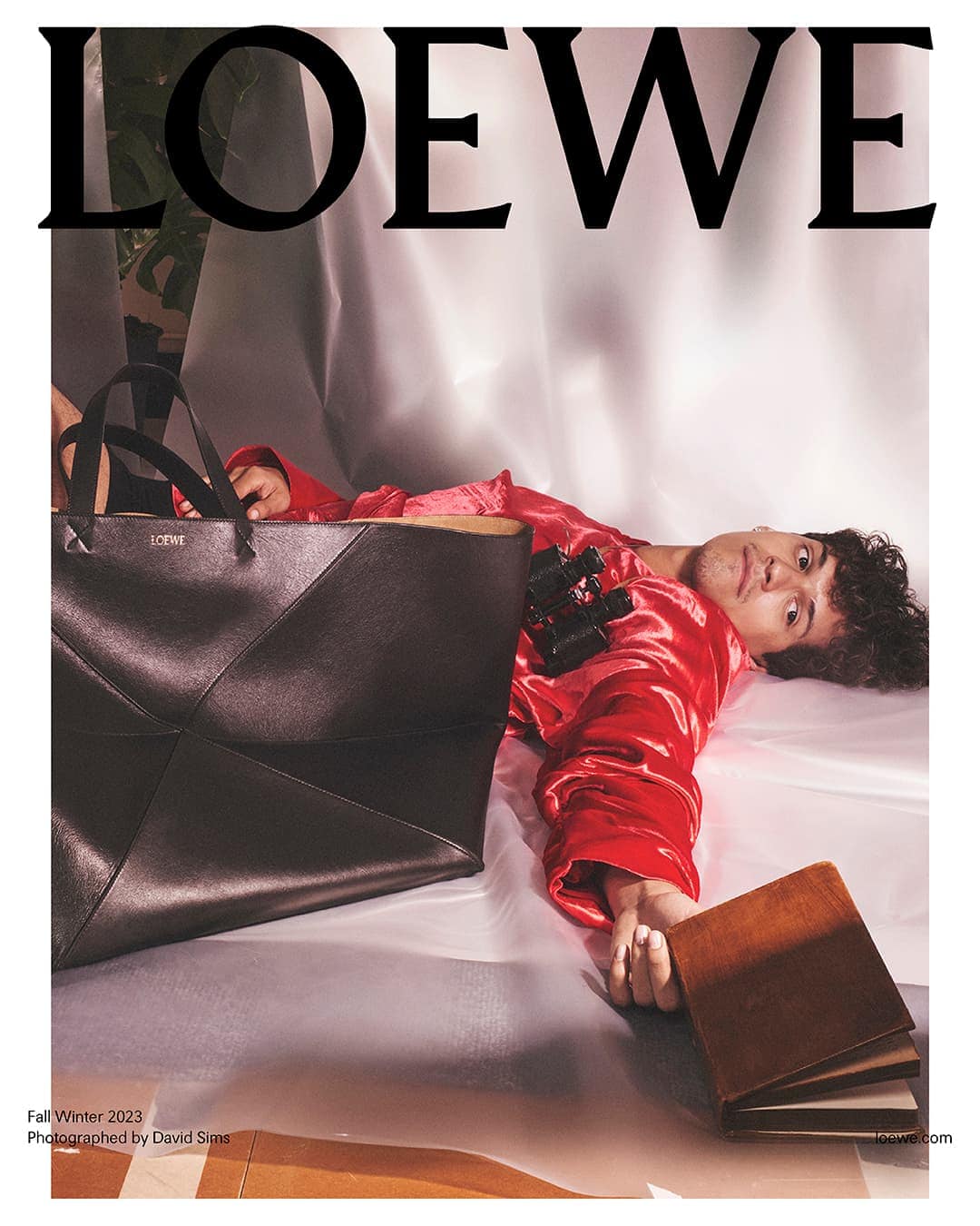 Fashion editorial featuring a model reclining with a black tote bag and book in a studio setting, showcasing Loewe's Fall/Winter collection.
