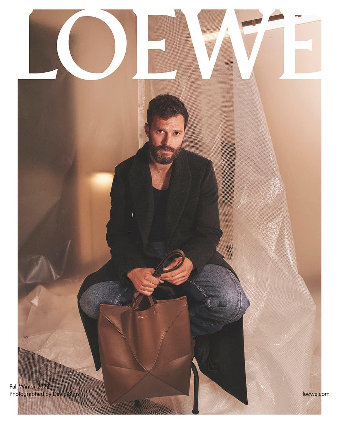 Jamie Dornan sits in a photo studio holding a brown leather tote bag in front of a textured white plastic sheeting backdrop.