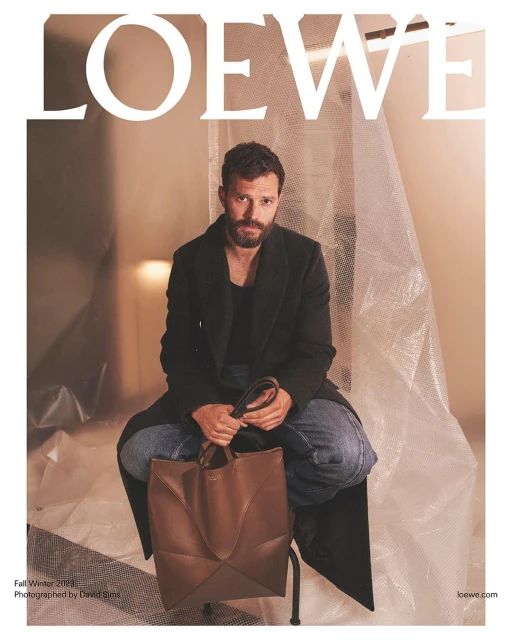 Jamie Dornan sits in a photo studio holding a brown leather tote bag in front of a textured white plastic sheeting backdrop.