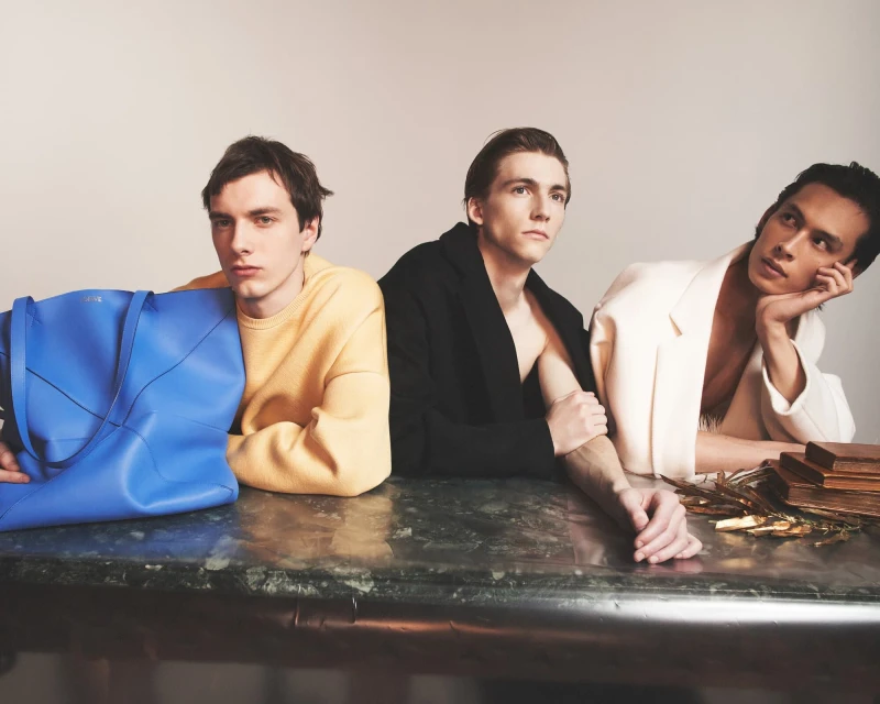 Three male fashion models pose at a marble table next to a large blue tote bag and gold foil in a brightly lit studio setting.