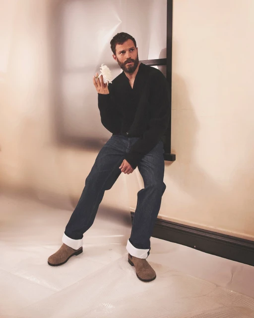 Man in jeans, black top, and boots poses indoors holding a small white object against a beige wall. He is standing near a window.