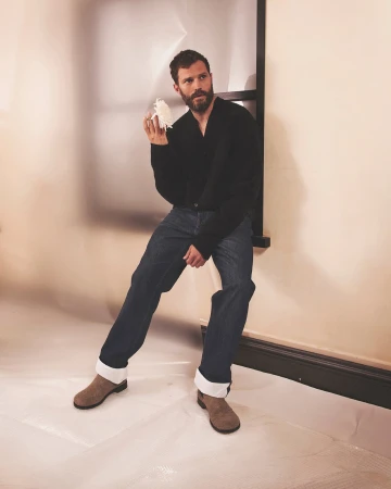 Jamie Dornan sitting against a set frame holding a white flower in a black knit sweater and cuffed denim for Loewe.
