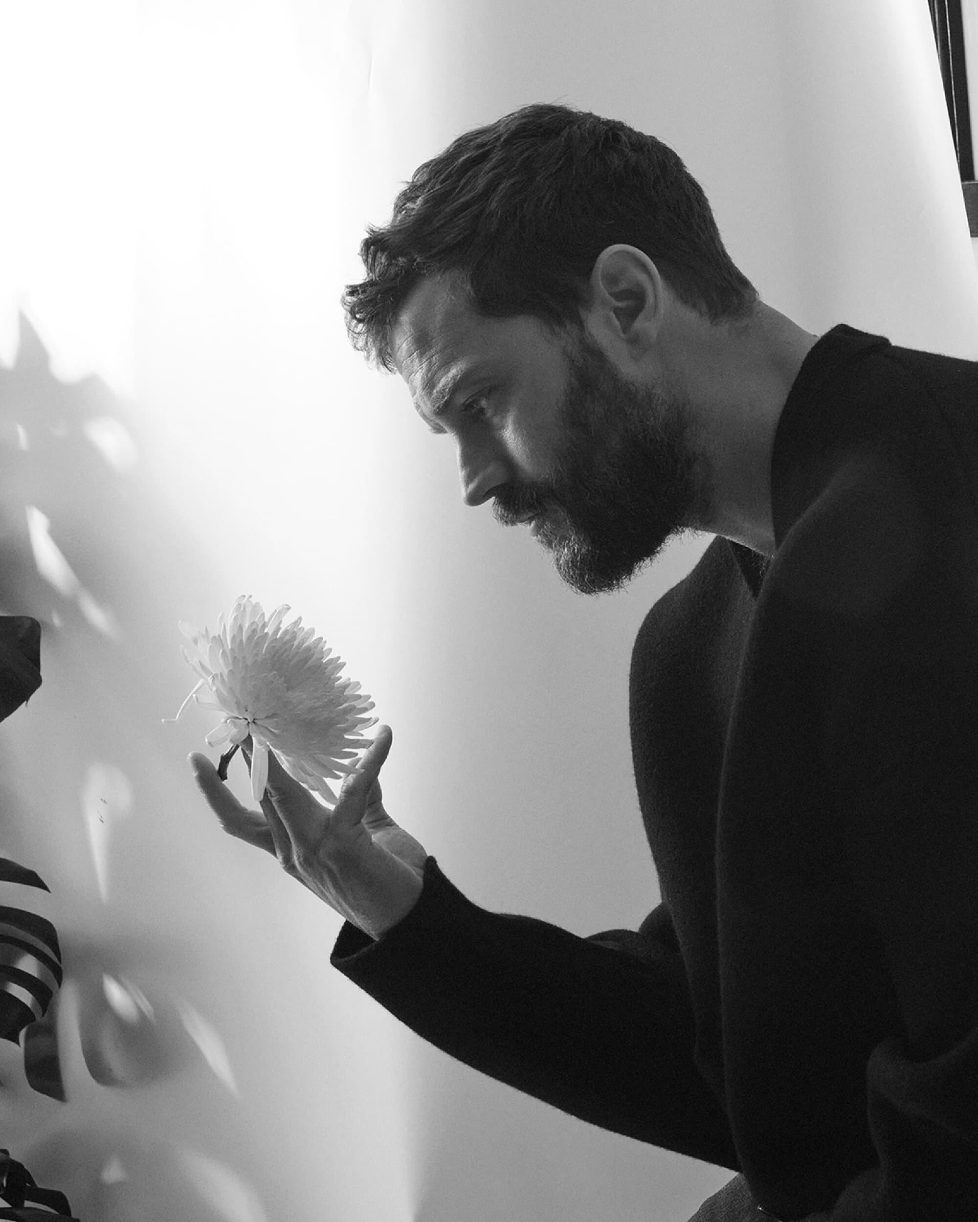Monochrome portrait of a bearded man in a dark sweater looking at a white flower held in his hand against a neutral background.