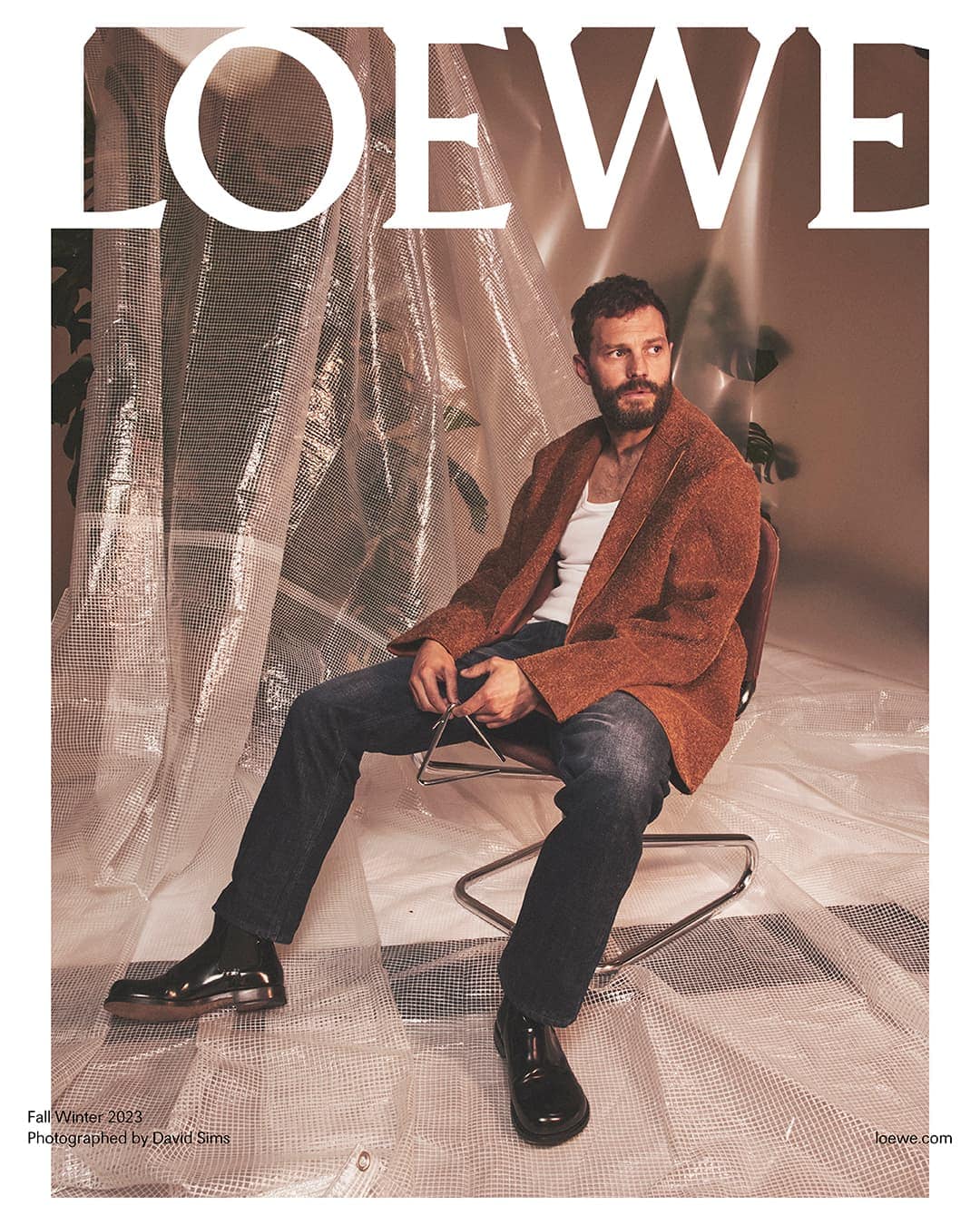 Fashion editorial featuring Jamie Dornan seated in a chair wearing a brown jacket, jeans, and black shoes in an indoor studio setting.