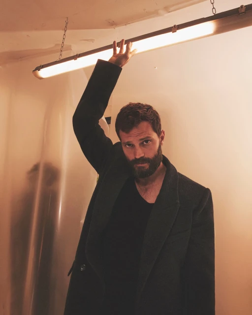 Bearded man in dark coat poses indoors with hand raised to fluorescent light, looking at the camera in medium shot. 