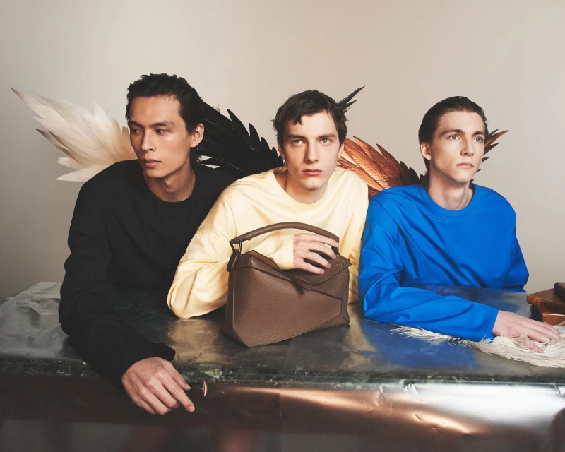 Three young adult models pose indoors with a Loewe Puzzle bag on a metallic surface, complemented by feather props against a neutral backdrop.