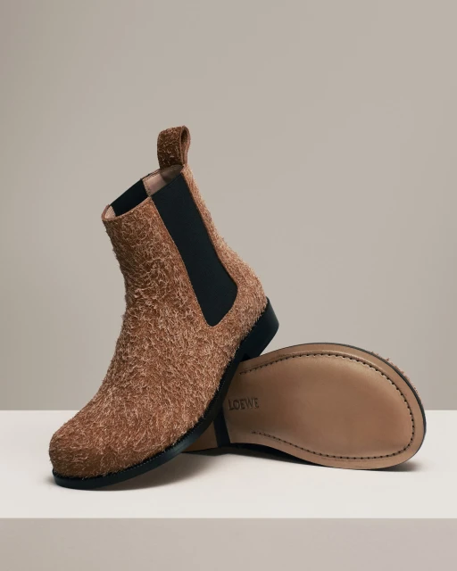 Studio shot of a pair of brown suede Loewe Chelsea boots, one upright and the other on its side displaying the sole.