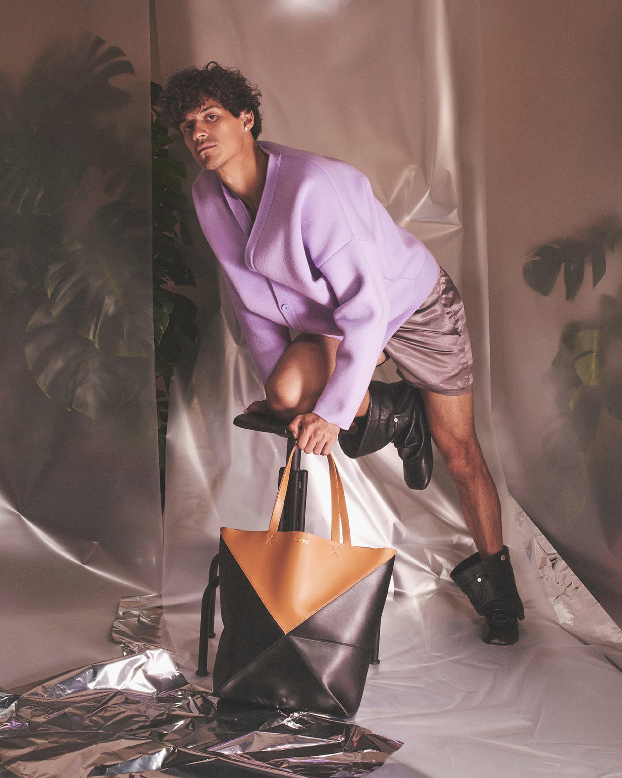 Fashionable adult male strikes a pose with a two-tone tote bag in a studio with silver reflective flooring and lavender color grading.