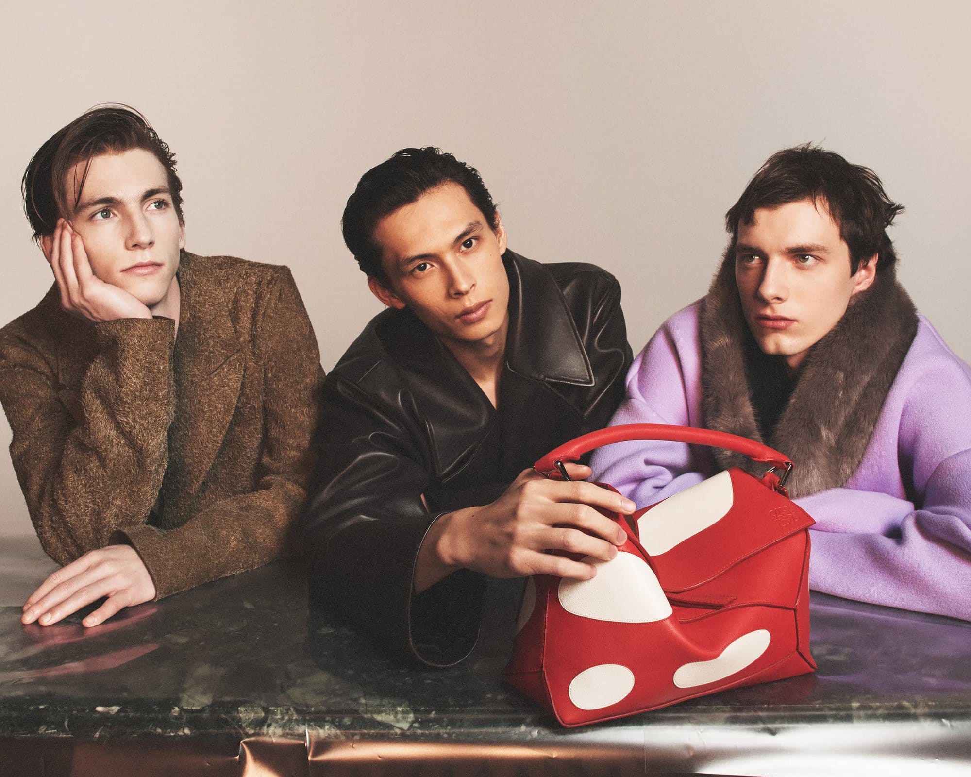 Three young male fashion models pose behind a table displaying a red and white handbag in a studio setting for a fashion editorial.