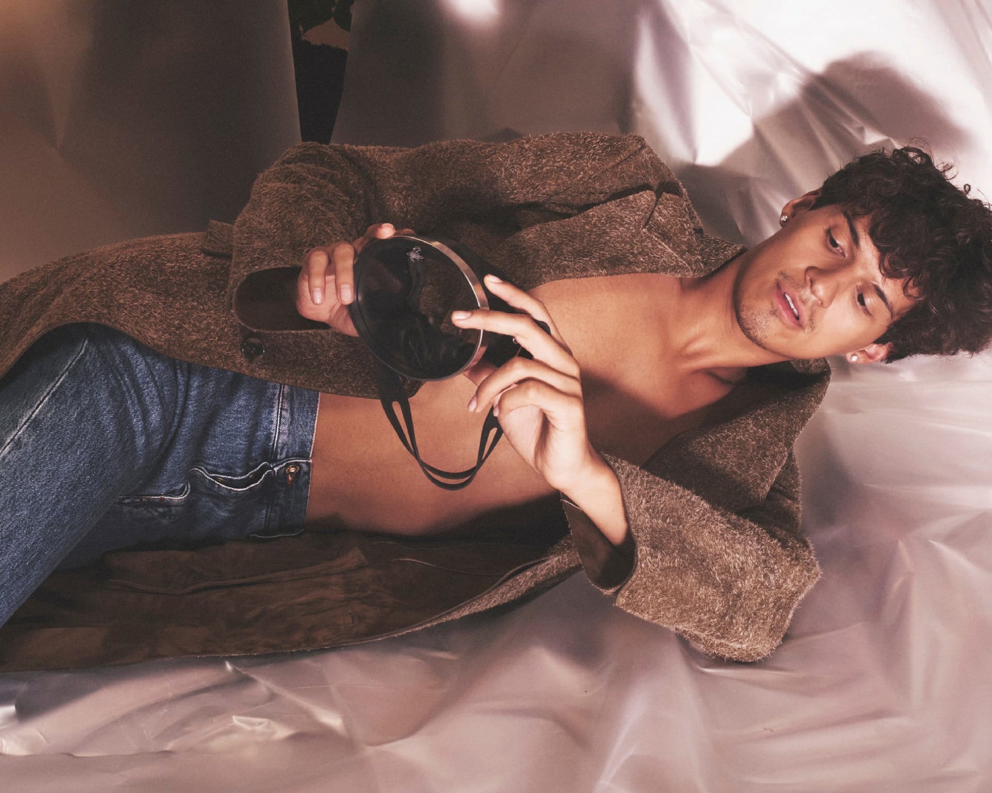 Young adult male poses with a compact mirror while wearing denim jeans and a brown jacket, laying on a white shiny surface.