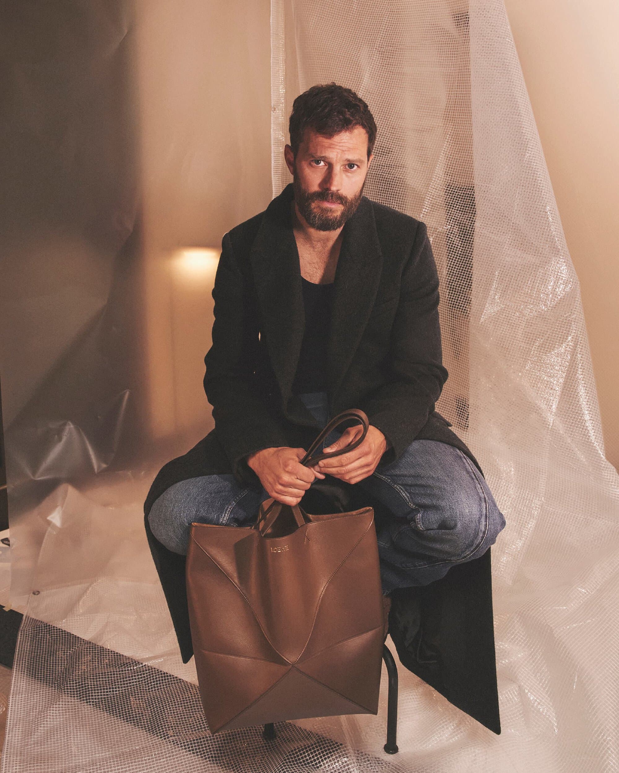 Stylish portrait of a handsome man in a coat, seated and holding a leather tote bag, against a textured backdrop. Fashion.