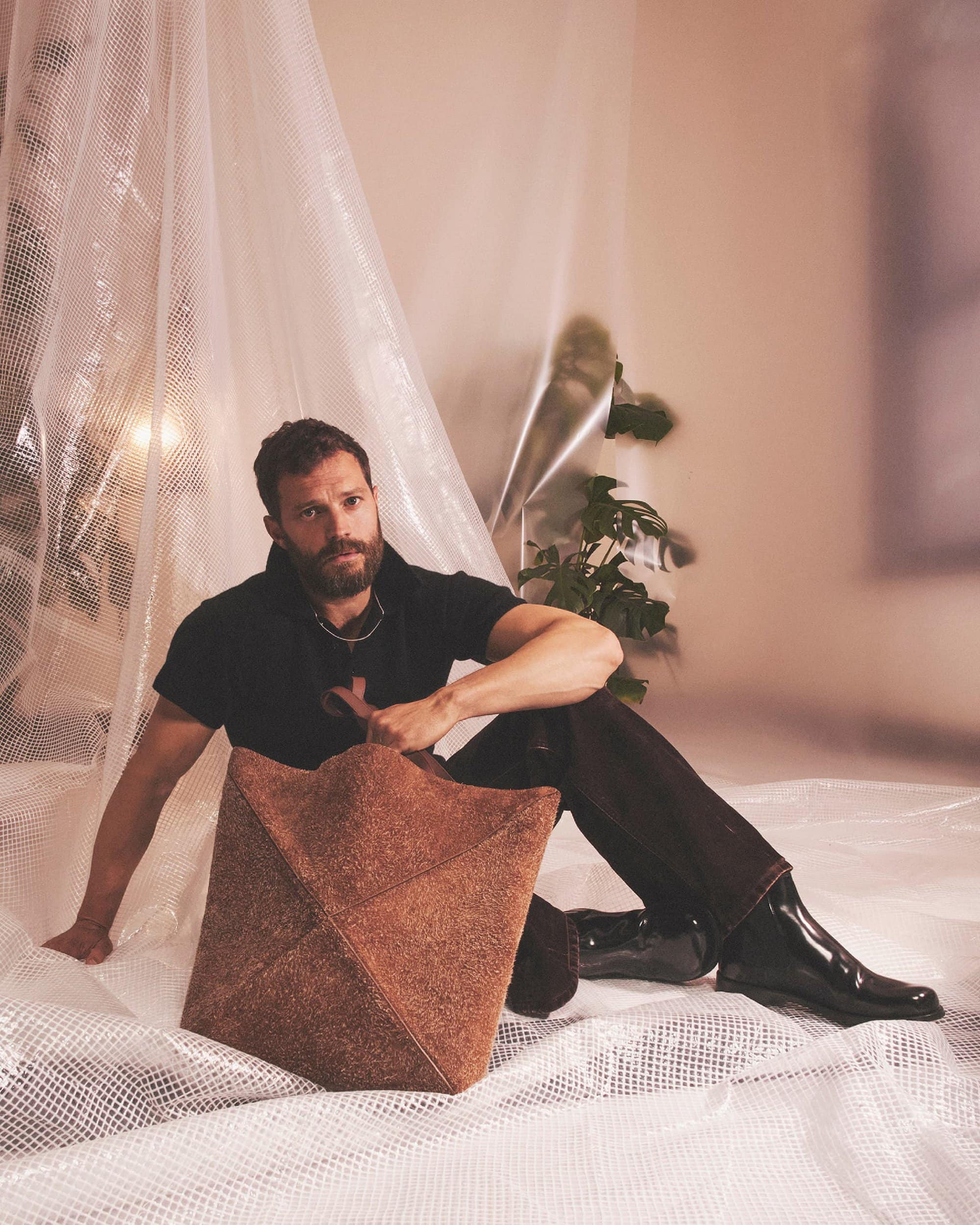 Actor Jamie Dornan poses seated holding a geometric tan bag in a photo studio with natural light, wearing a black shirt and black boots.