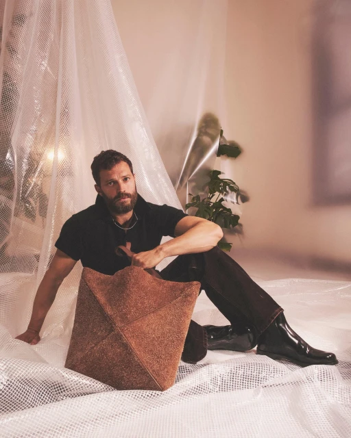 Actor Jamie Dornan poses seated holding a geometric tan bag in a photo studio with natural light, wearing a black shirt and black boots.