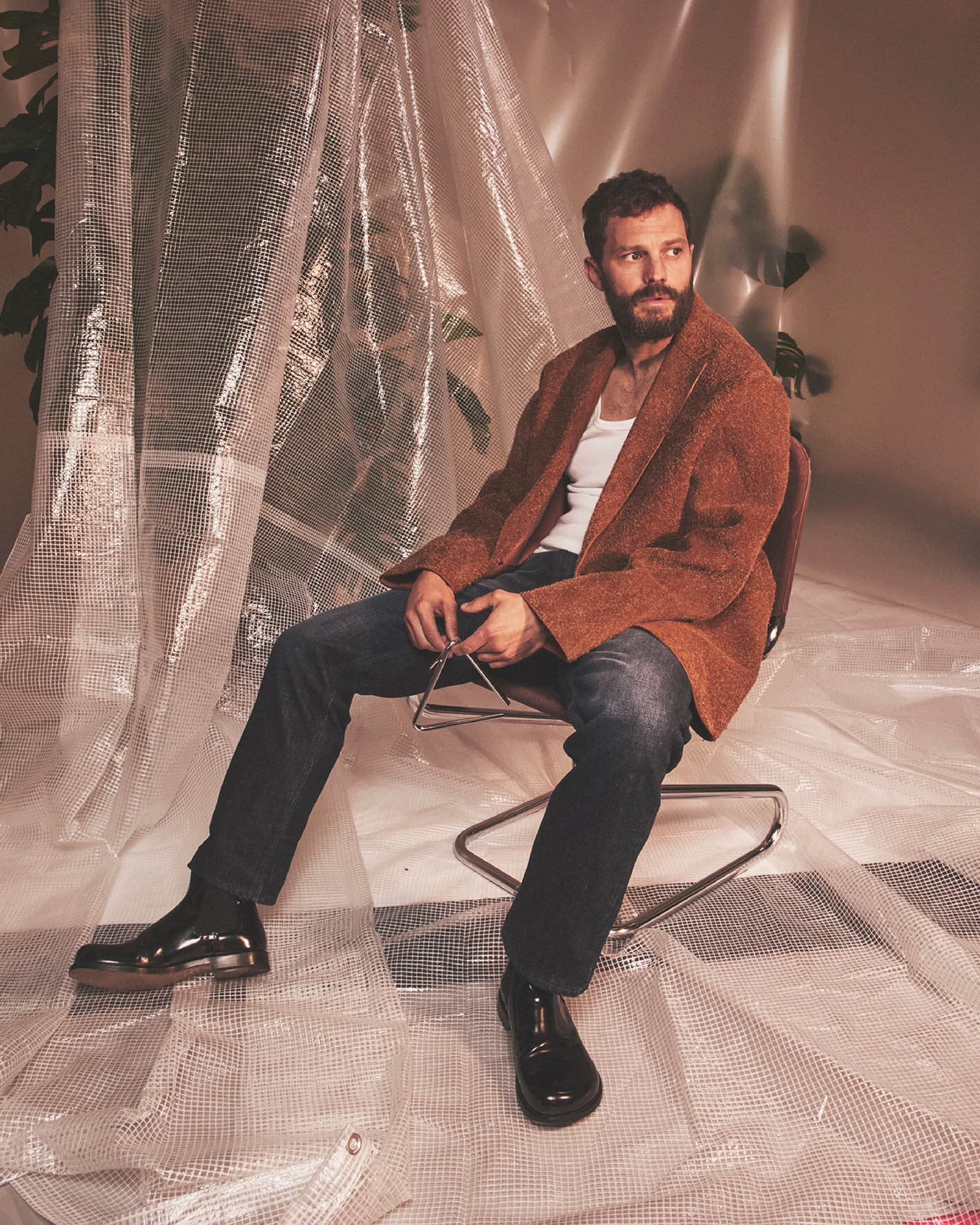 Man with a beard sitting on a chair, wearing a brown jacket, white shirt, and jeans, posing in front of a clear bubble-wrap backdrop.