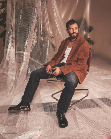 Man with a beard sitting on a chair, wearing a brown jacket, white shirt, and jeans, posing in front of a clear bubble-wrap backdrop.