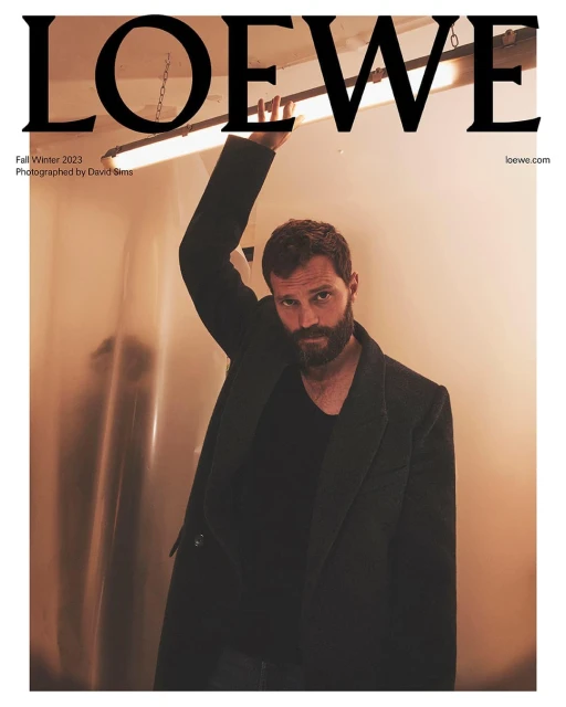 Man with beard posing indoors wearing a dark coat over a black shirt in a fashion magazine cover shot for Loewe Fall/Winter 2023.
