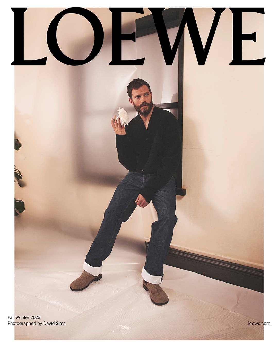 Jamie Dornan poses in Loewe Fall/Winter 2023 ad, wearing a black cardigan, jeans, and suede boots, with the Loewe logo.