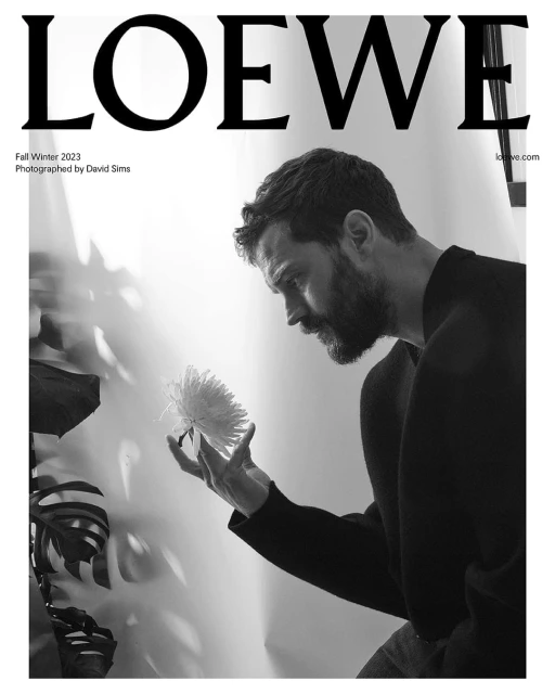 Black-and-white portrait of a bearded man in a dark shirt gently holding a white dahlia flower near a tropical plant, indoor.