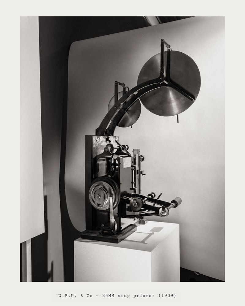 Vintage 35mm step printer on a white cube against a neutral background. The printer is metallic, with spools and intricate mechanisms.