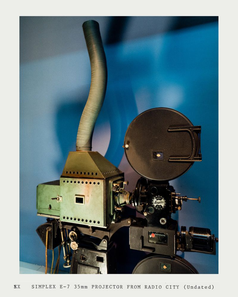 Vintage Simplex E-7 35mm film projector, Radio City model, featuring aged metal, coiled exhaust, and a bright blue backdrop.