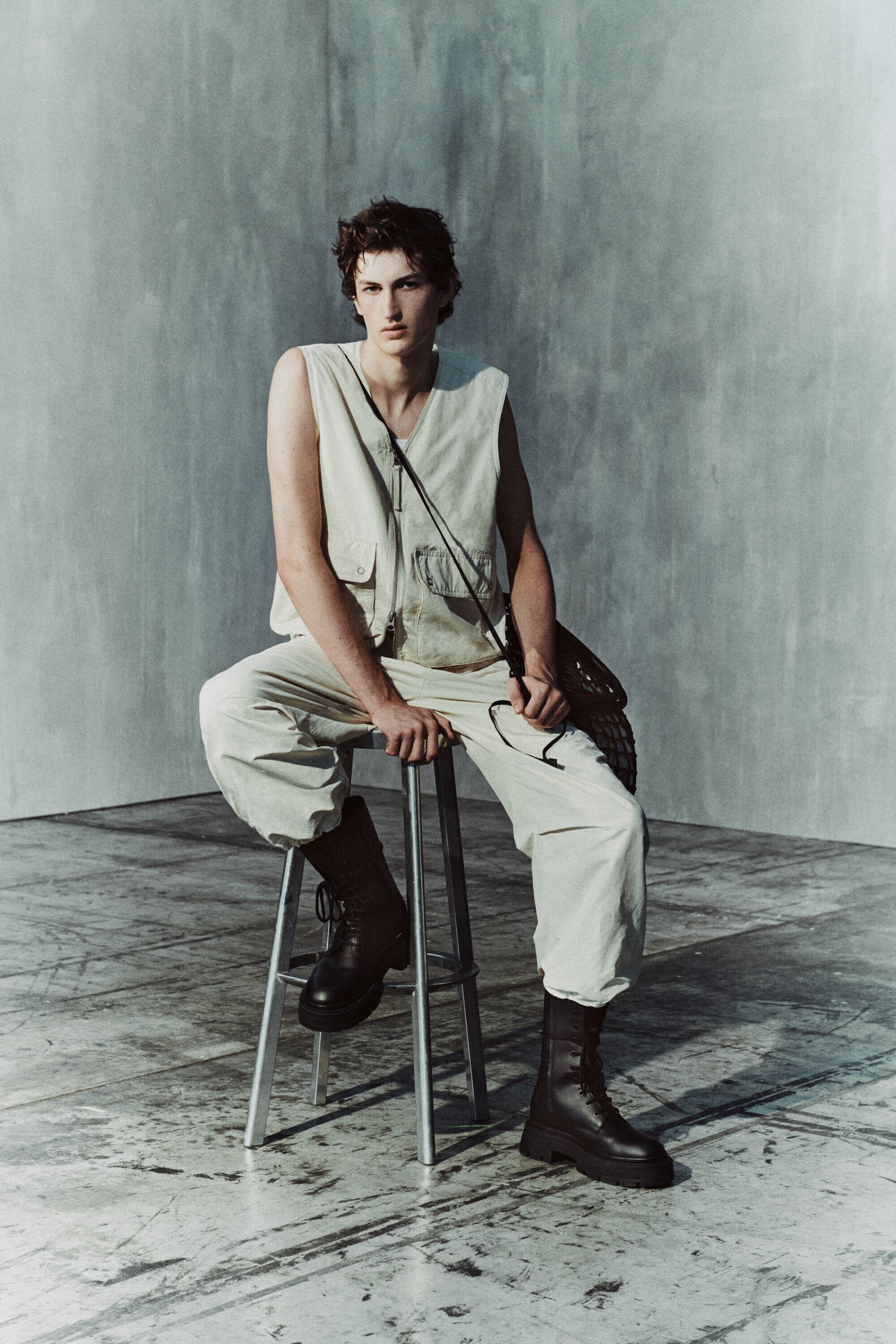 Young man in a utilitarian outfit sits on a stool in a photo studio with a grey backdrop, wearing boots and a shoulder bag. 