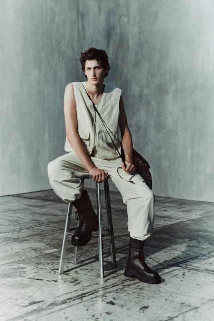 Young man in a utilitarian outfit sits on a stool in a photo studio with a grey backdrop, wearing boots and a shoulder bag. 