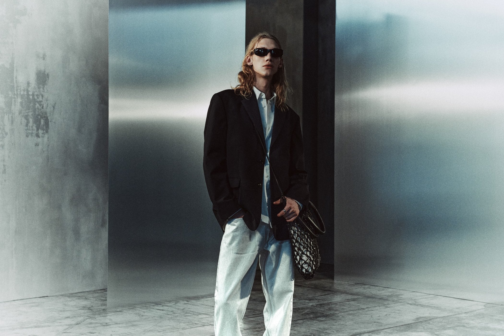 Adult male with long hair and sunglasses in a black jacket, white shirt, and light blue jeans stands against a modern gray background.