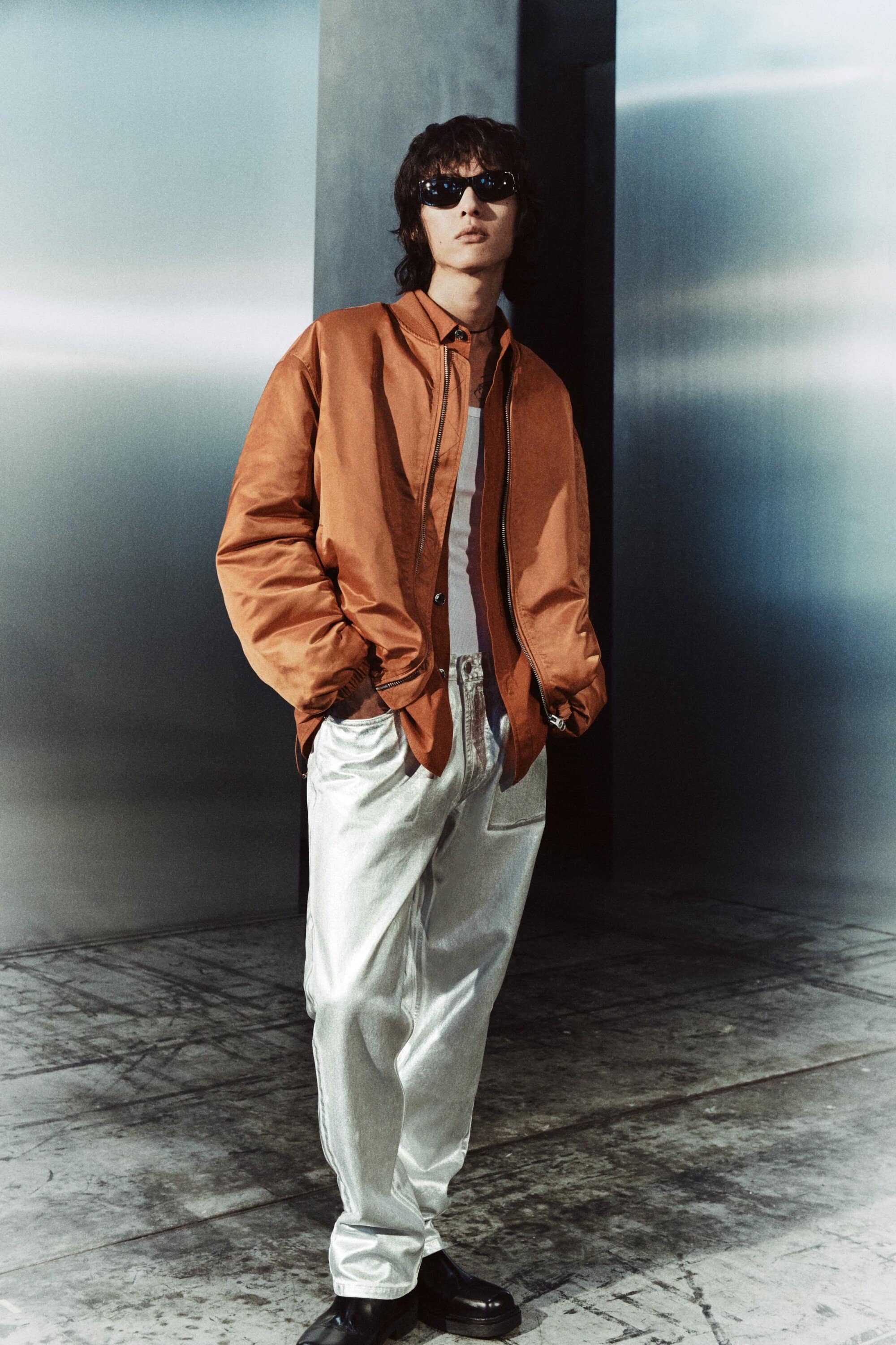 Stylish man in sunglasses and an orange jacket poses in a studio with metallic walls and floors, showcasing a fashion-forward look.