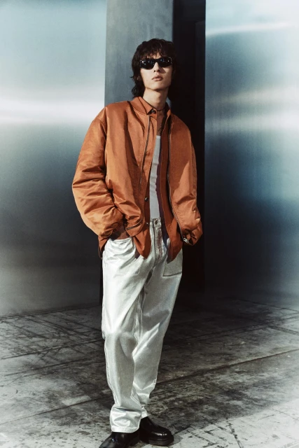 Stylish man in sunglasses and an orange jacket poses in a studio with metallic walls and floors, showcasing a fashion-forward look.