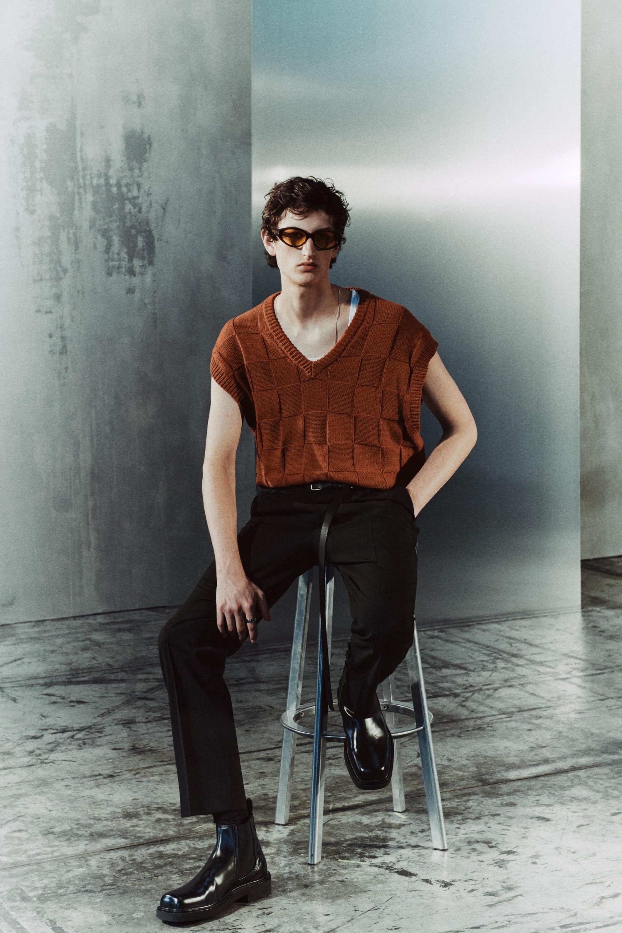 A young adult man is seated on a metal stool wearing sunglasses, a brown knit vest, black trousers, and black Chelsea boots, with a grunge-style backdrop.