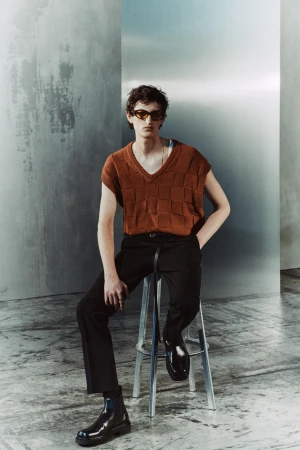 A young adult man is seated on a metal stool wearing sunglasses, a brown knit vest, black trousers, and black Chelsea boots, with a grunge-style backdrop.