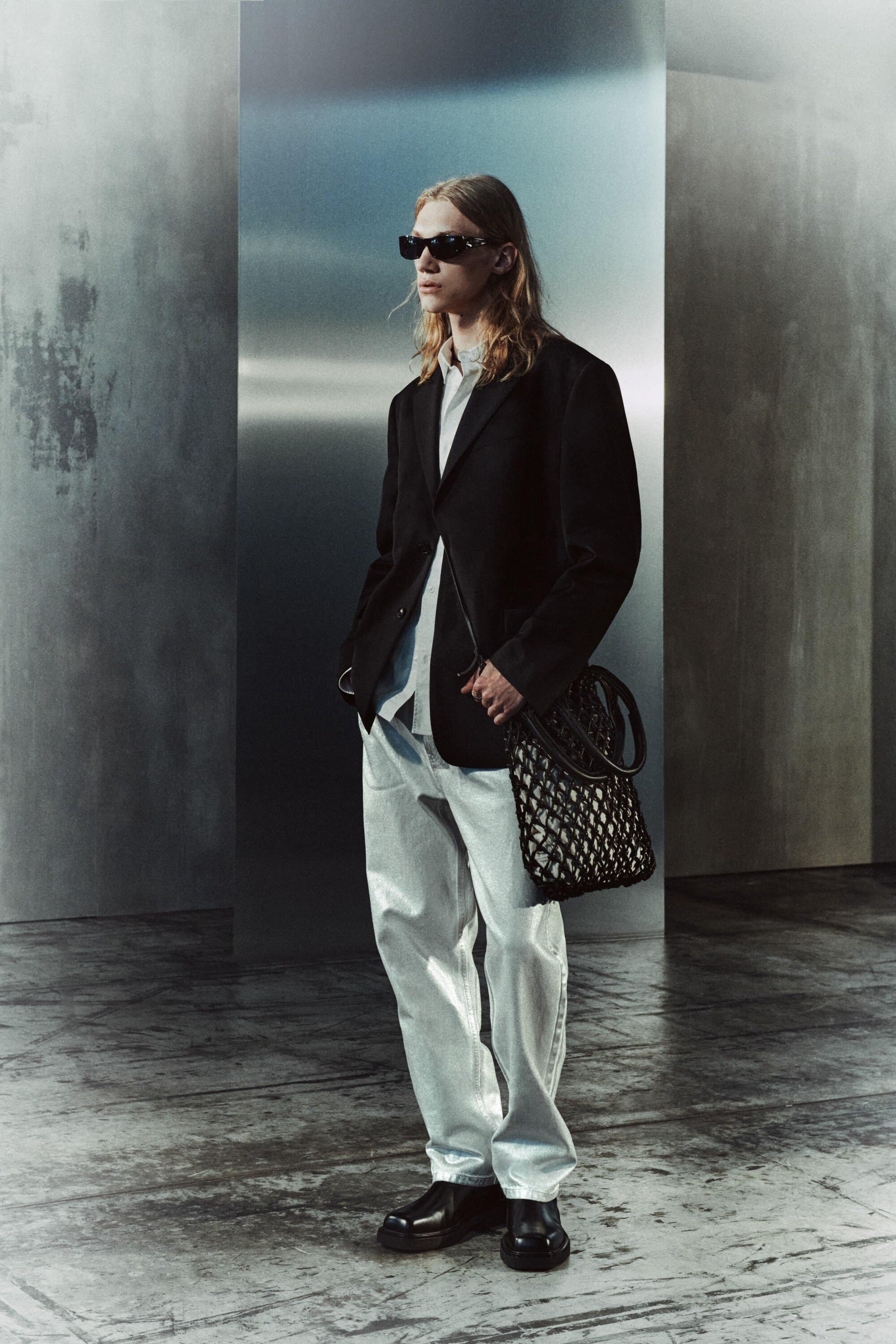 Man in blazer, light jeans, sunglasses, and black loafers poses in a silver-grey studio with a mesh bag. Fashion editorial.