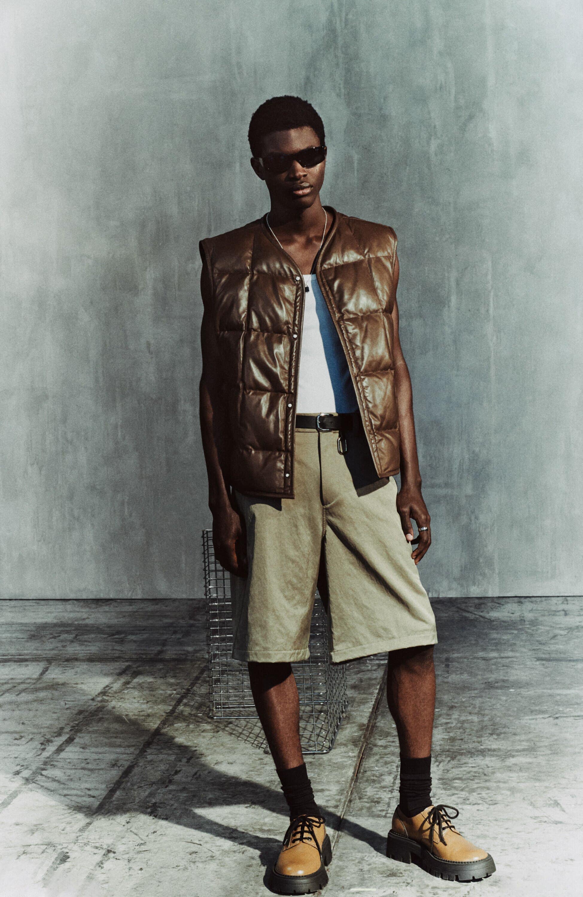 Dark-skinned male model in a brown puffer vest, white t-shirt, and khaki shorts, standing against a concrete wall in a fashion editorial shoot.