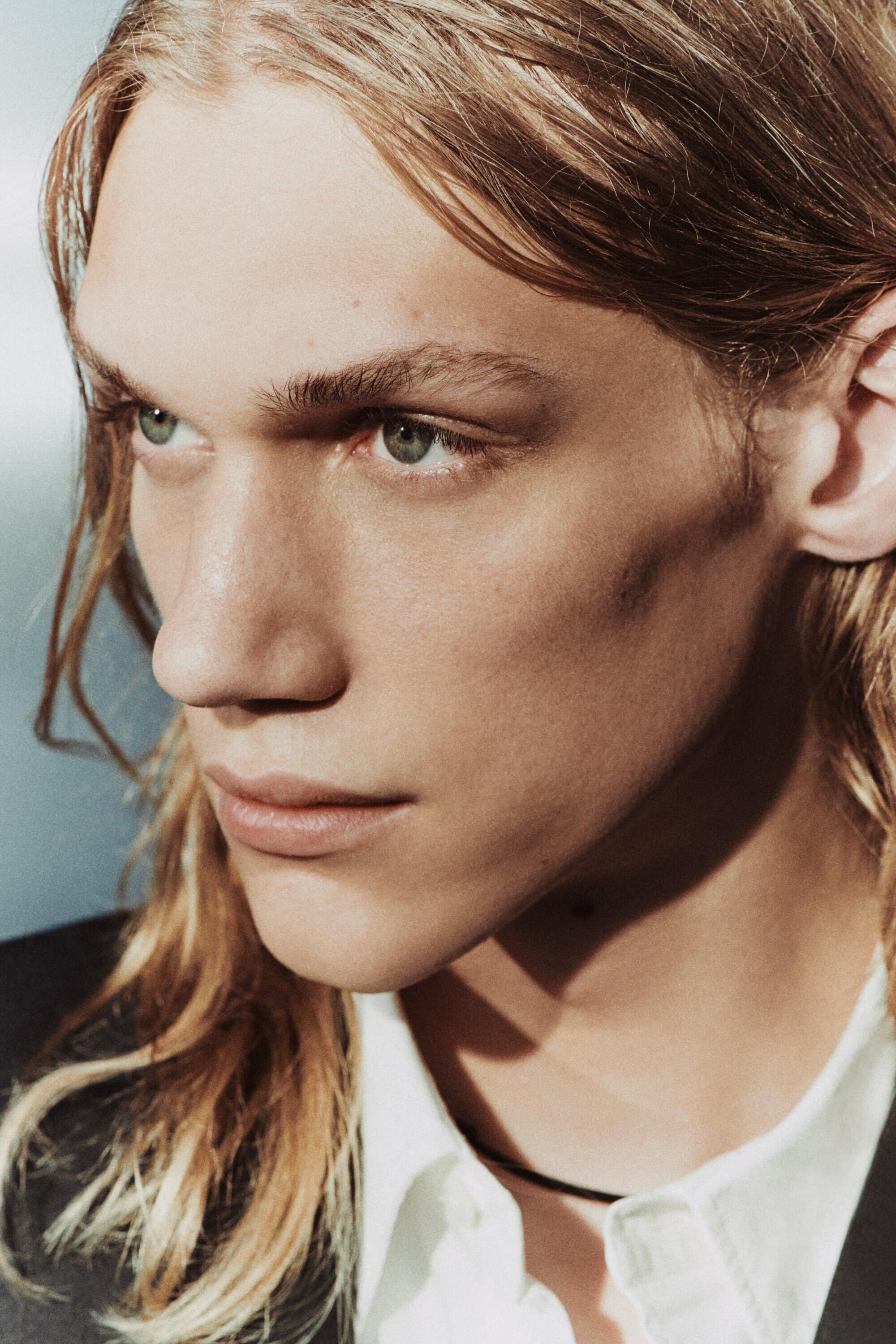Close-up studio portrait of a fair-skinned adult male with long blond hair, wearing a white shirt and black jacket, against a light blue background.