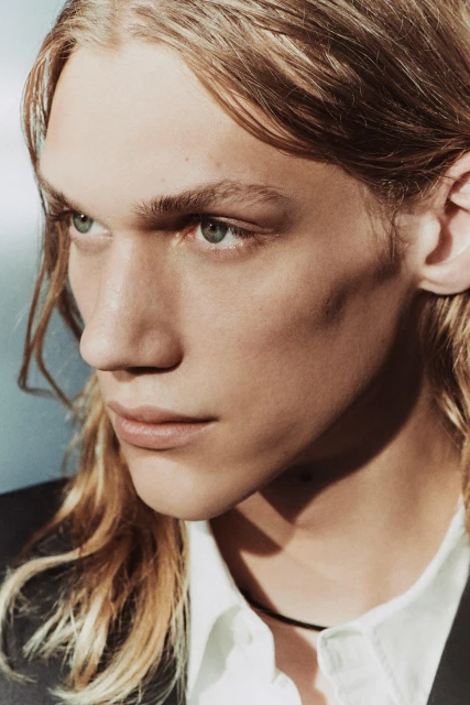 Close-up studio portrait of a fair-skinned adult male with long blond hair, wearing a white shirt and black jacket, against a light blue background.