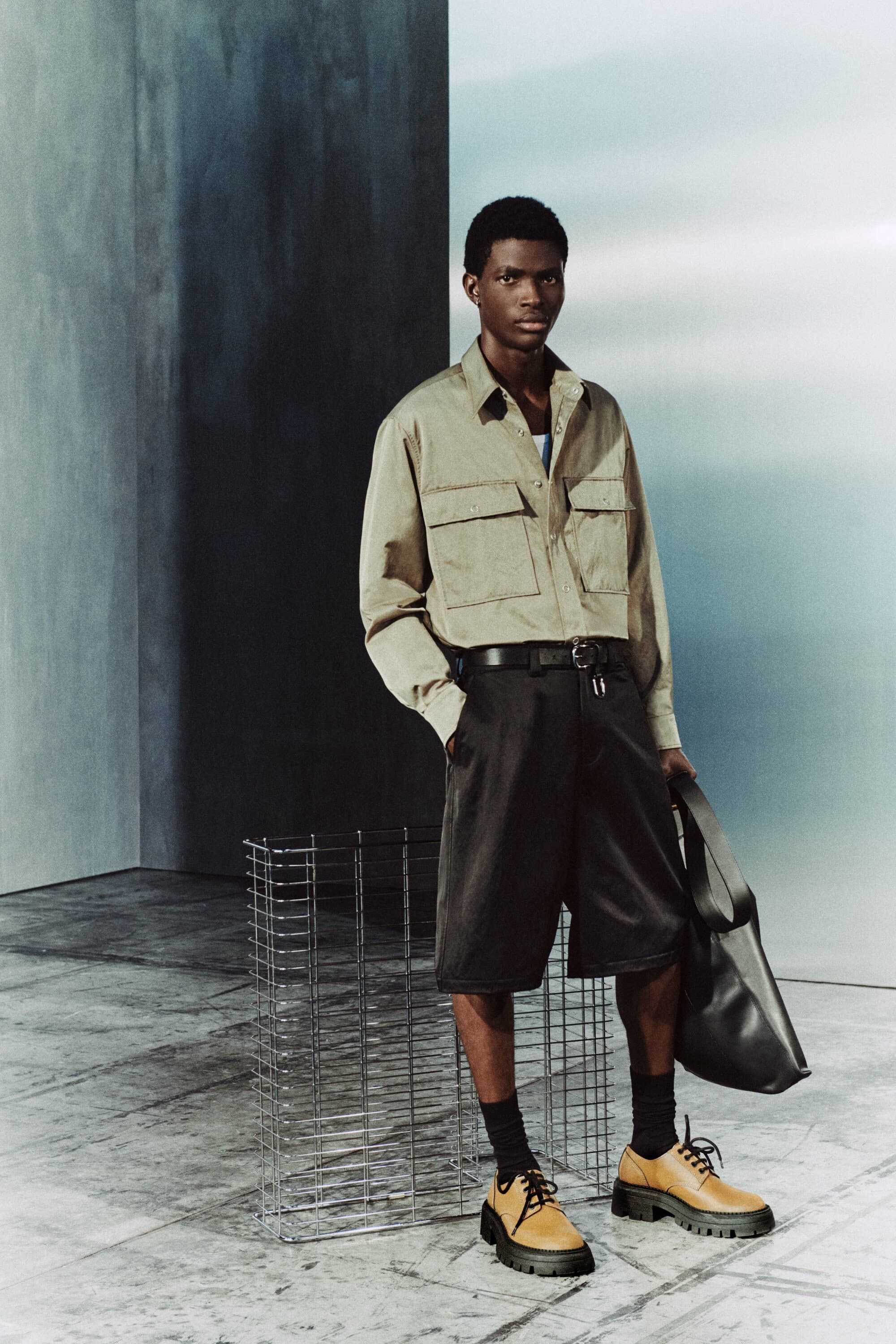 Dark-skinned male model wears a light-brown button-up shirt, dark shorts, black socks, tan shoes, and carries a black bag in a studio setting.