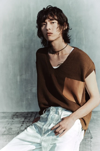 A young Asian man is posing in a fashion shot, wearing a brown sleeveless sweater and light jeans, with a muted blue studio background.