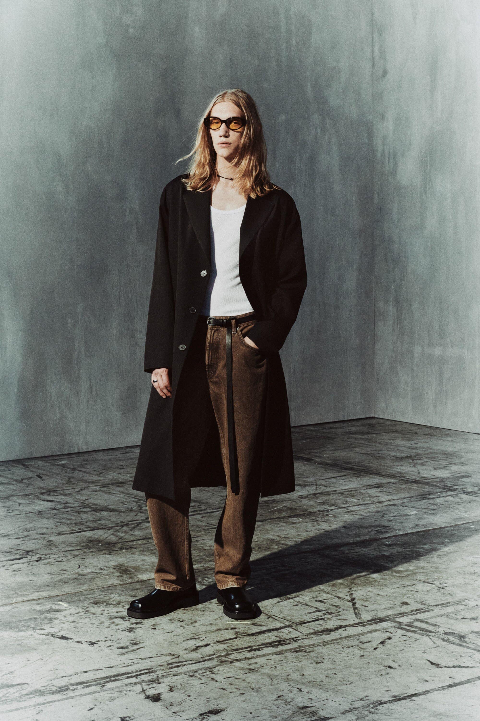 Full shot of androgynous person wearing a black overcoat, white tank top, brown pants, and black shoes in a studio setting.