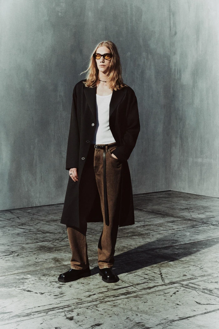 Model with long blonde hair in sunglasses, wearing an open black overcoat, white tank top, and brown trousers on a concrete floor.