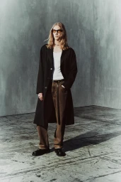 Model with long blonde hair in sunglasses, wearing an open black overcoat, white tank top, and brown trousers on a concrete floor.