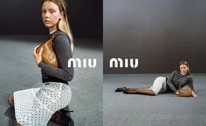Mia Goth holding a brown leather bag wearing a grey cardigan and polka dot skirt against a grey seamless backdrop