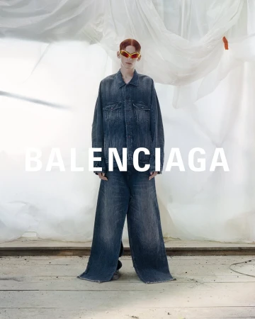 Fashion shot of a person with red hair wearing orange sunglasses, denim jacket and pants, standing in a photo studio in front of white cloth.