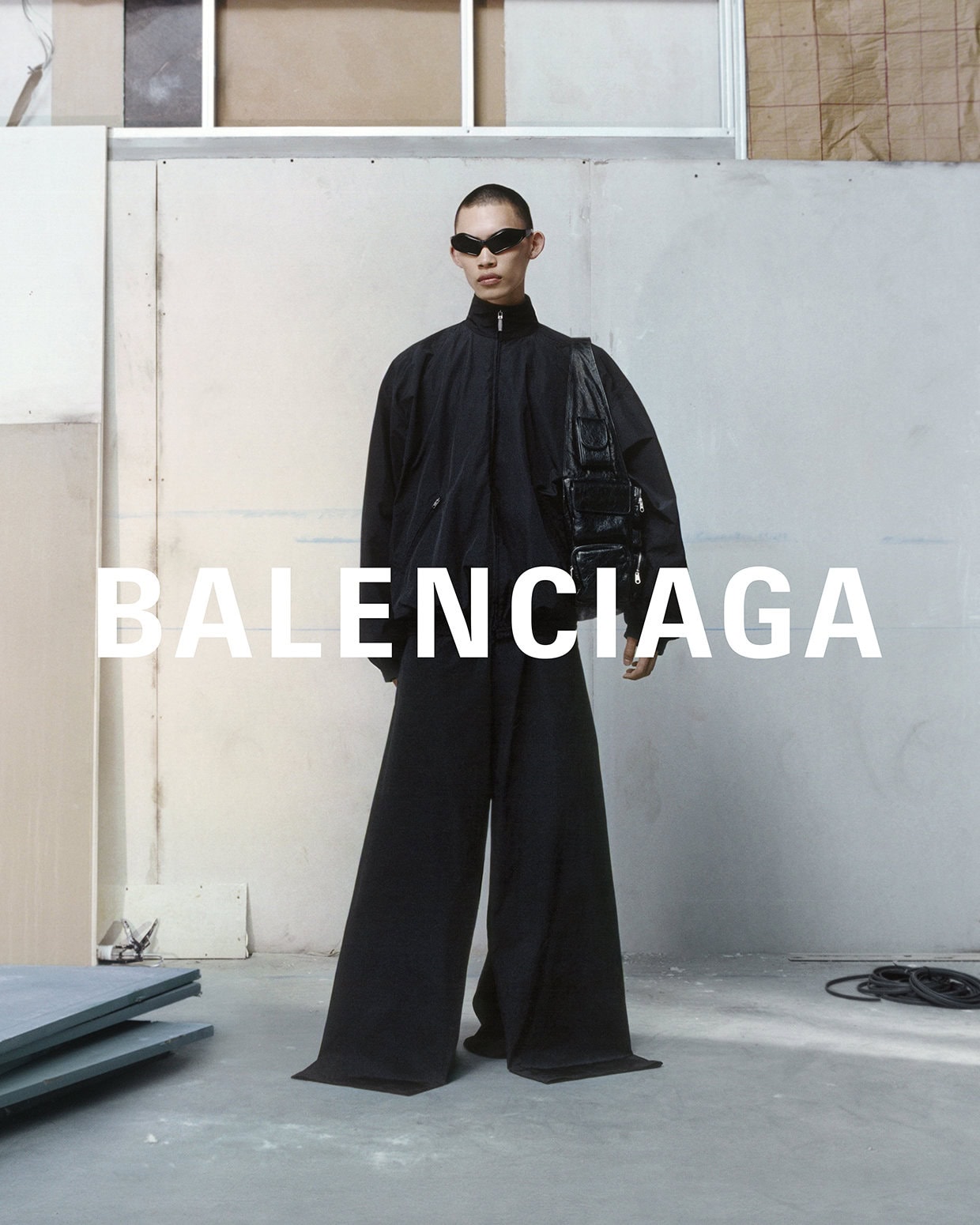 A person in sunglasses and black clothing poses indoors in front of the brand name BALENCIAGA. They carry a black bag.