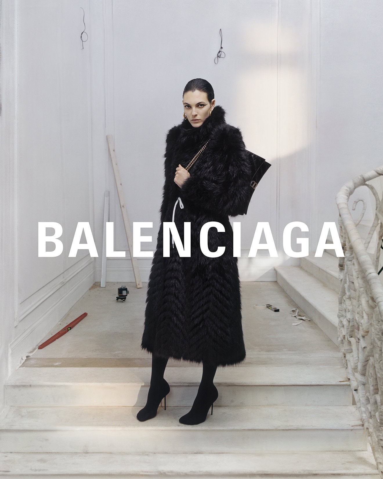 Fashion editorial featuring a woman in a black faux fur coat holding a handbag, posing on stairs in a minimalist white interior.