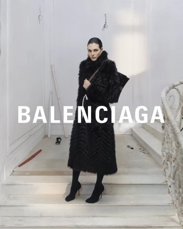 Fashion editorial featuring a woman in a black faux fur coat holding a handbag, posing on stairs in a minimalist white interior.
