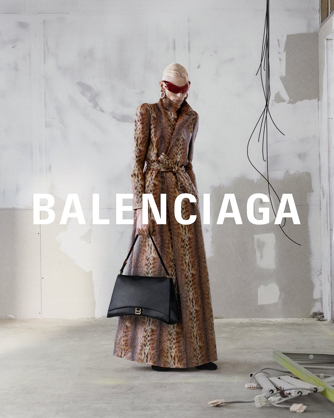 A blond model poses in a full-length animal print coat and black Balenciaga handbag against a textured gray studio wall for a fashion editorial.