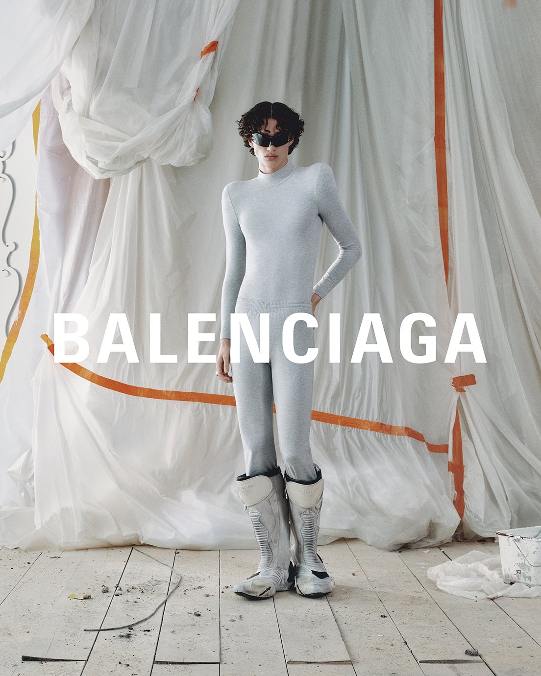 Fashion model in a light grey long-sleeved shirt, pants, and silver boots poses in front of draped white fabric with orange accents.
