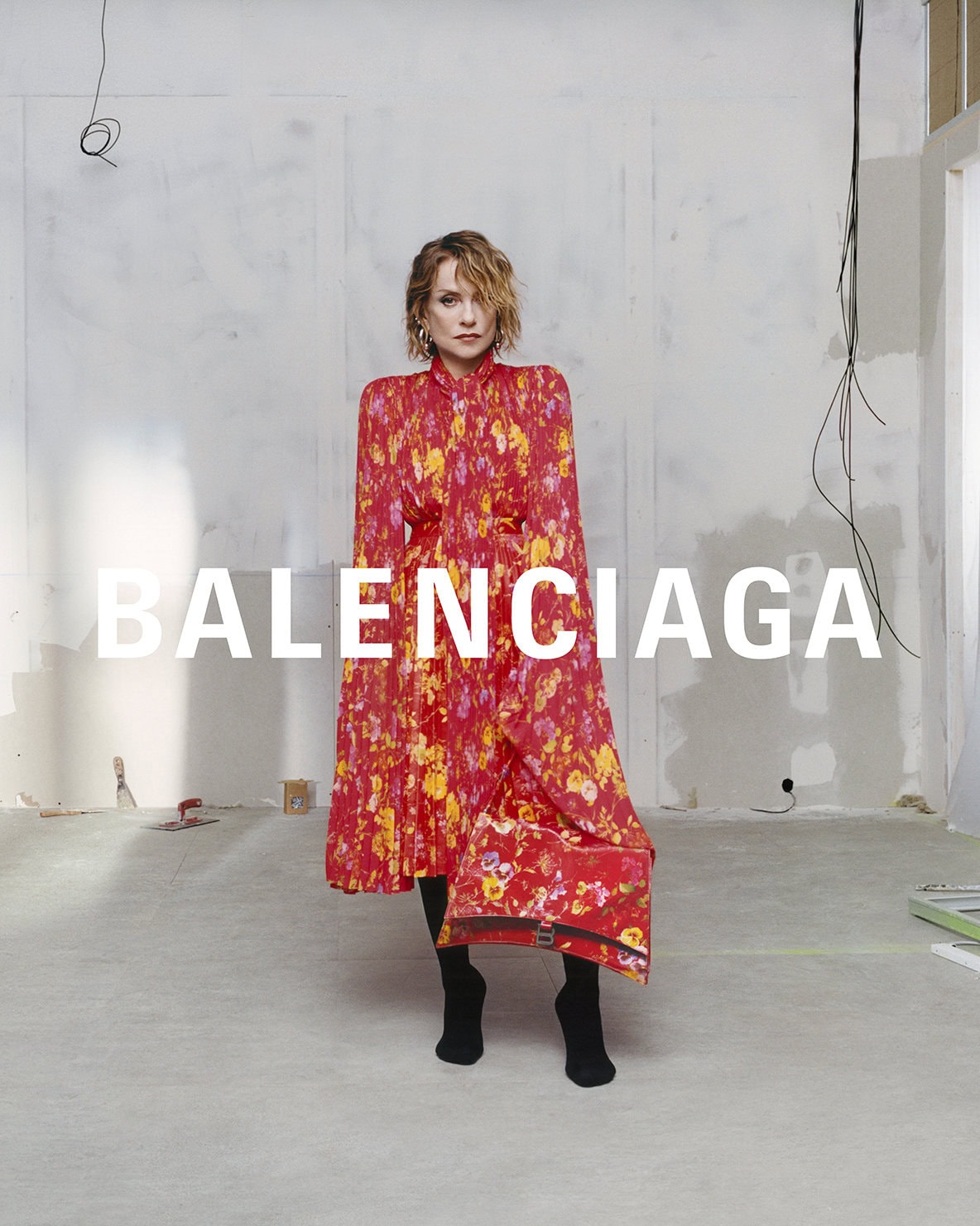Woman posing in a red floral dress with a matching purse in a studio with unfinished walls, advertising Balenciaga fashion brand.